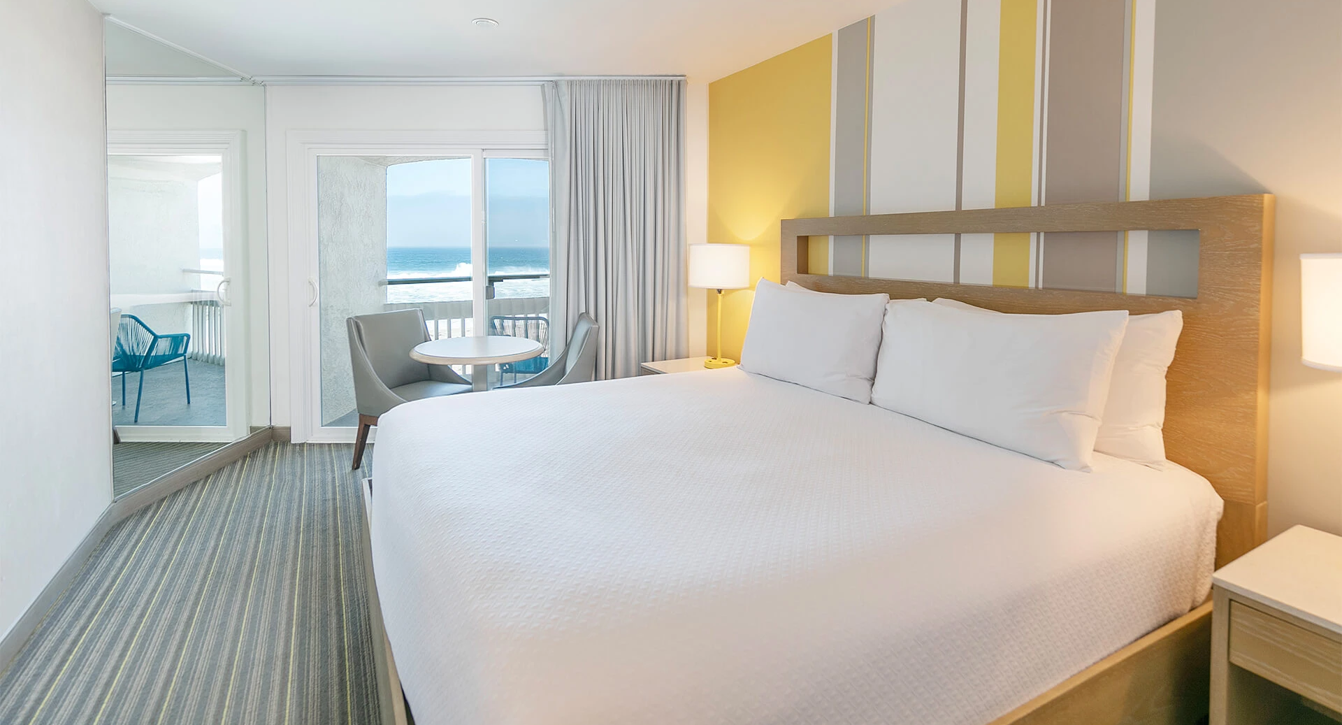 A bright hotel room with a large white bed, two nightstands, a desk and chairs, and a balcony with a sea view beyond sheer curtains.