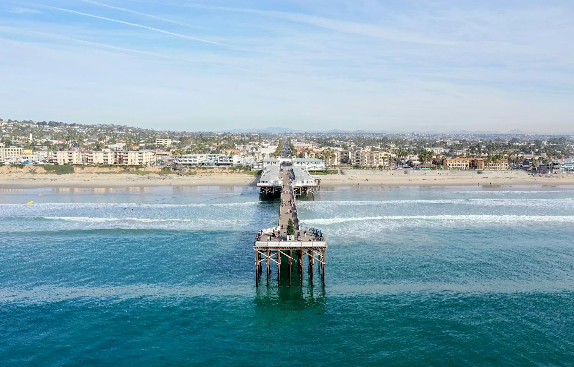 A long pier extends into blue water toward a distant sandy beach with buildings along the shore, under a clear sky.