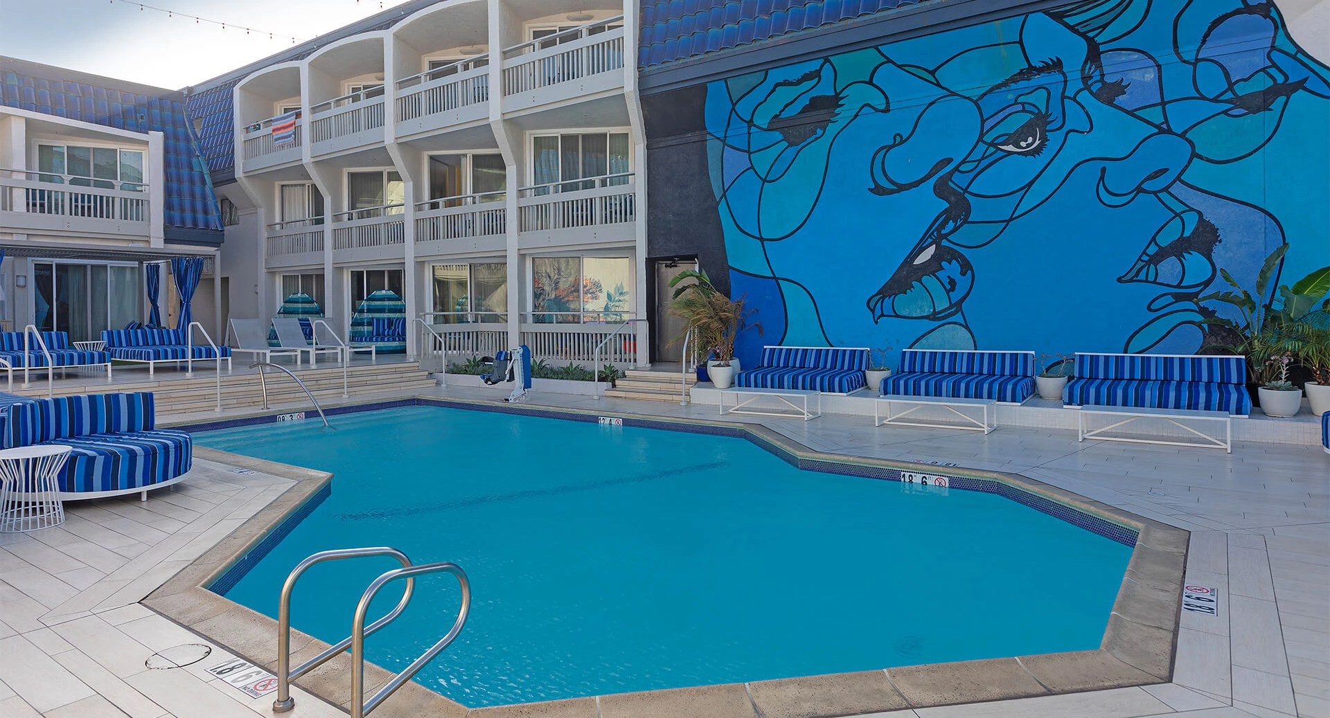 A hotel pool area with blue loungers and a striking blue mural on the wall, surrounded by white balconies and sunny skies. Ending sentence.
