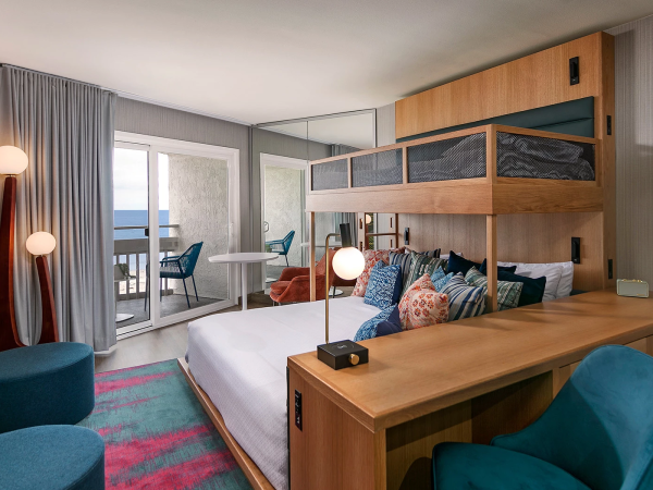 A bright hotel room with a bed and bunk, wooden desk, teal chairs, a balcony, and soft lighting. Ends with a period.