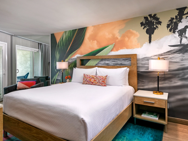 A modern hotel bedroom with a mural wall featuring palm trees and a beach, a wooden bed frame with white bedding, nightstands, lamps, and a seating area with colorful chairs.