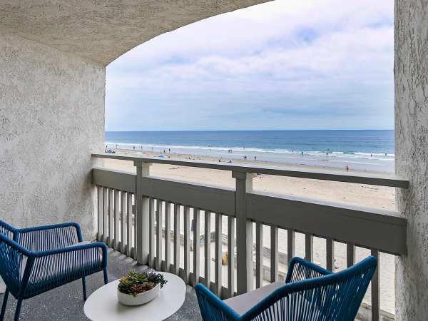A balcony view of a sandy beach and blue ocean, with two blue chairs and a small round table holding a plant, overlooking the shore.