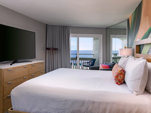 A modern hotel room with a large bed, flatscreen TV, wooden dresser, and a balcony overlooking the ocean, bright and inviting.