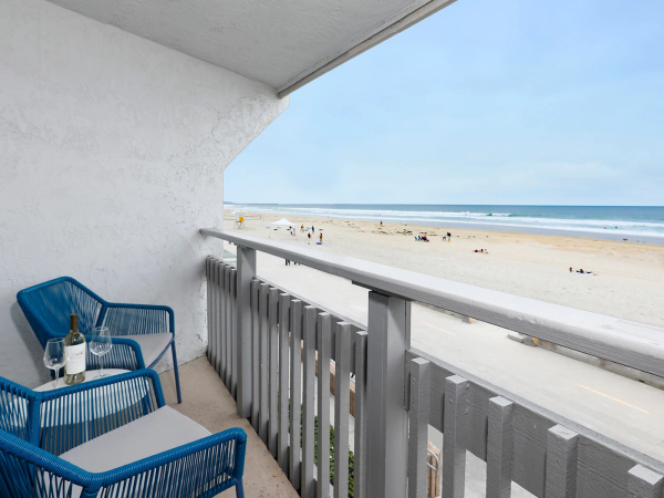 A balcony with blue wicker chairs and a small table overlooking a sunny beach and calm ocean, people sunbathing on the sandy shore.