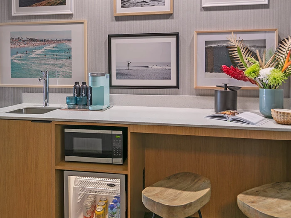 A compact kitchenette with a sink, mini fridge, microwave, and a wooden counter; framed beach photos on the wall and two stools.