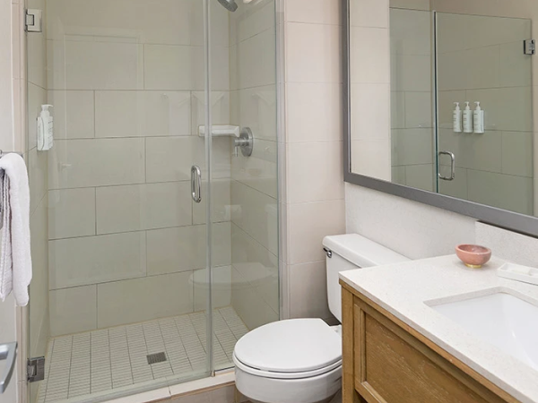 A modern bathroom with a glass-enclosed shower, wall-mounted toilet, and a wooden vanity with a white countertop and mirror above, next to a towel rack.