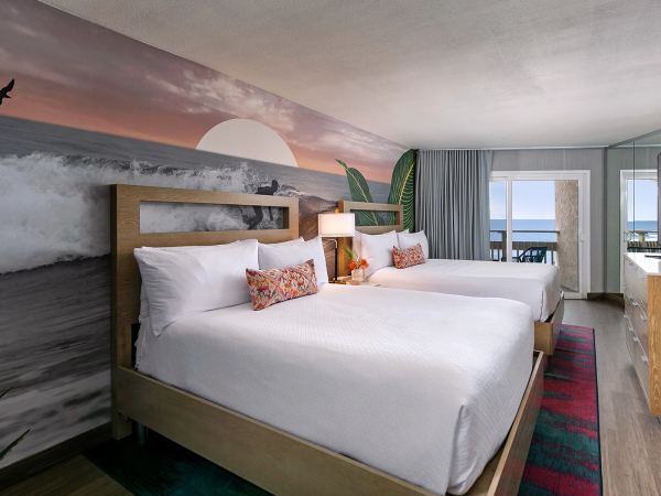 A hotel room with two queen beds, coastal wall mural of sunset waves, bright decor, desk, flat-screen TV, and a sea view through the window.