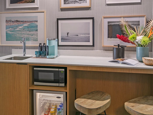 A compact kitchenette with a sink, microwave, mini fridge, and framed beach photos on the wall.