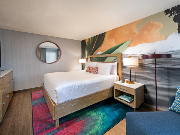 A stylish hotel room with a king bed, colorful rug, wall mural of waves, round mirror, nightstands, lamp, and a blue sofa.