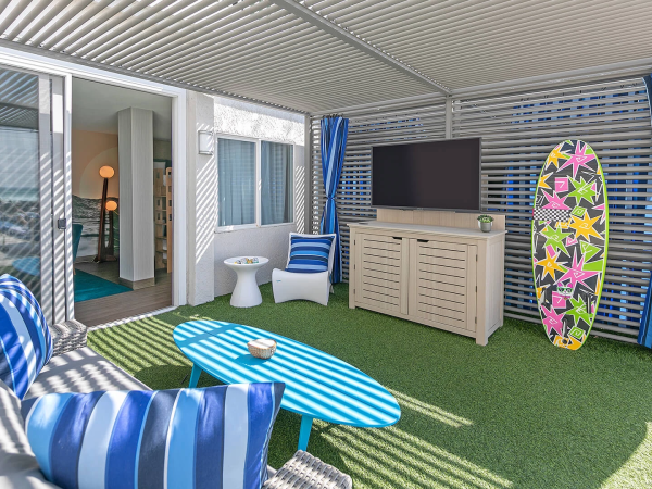 A sunny covered patio with striped blue cushions, a turquoise table, artificial grass, a flat-screen TV, a dresser, surfboard, and blue curtains.