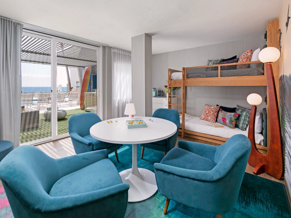 A cozy hotel room with a round white table, four teal chairs, a bunk bed, sea wall mural, and large sliding doors opening to a balcony.