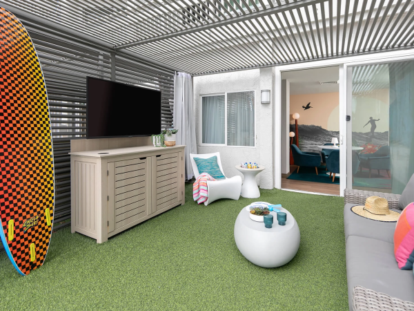 A stylish rooftop lounge with artificial grass, a surfboard, outdoor TV, wicker cabinet, cozy seating, ottoman, bright cushions, and sliding doors to indoor space.