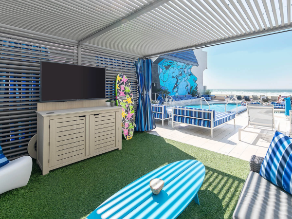 A stylish outdoor cabana with blue-striped cushions, a TV on a cabinet, a surfboard, turf rug, and a sunny sea-view lounging area.