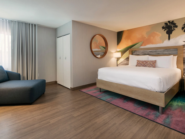 A modern hotel room with a king bed, colorful mural wall, nightstands, a cozy sitting area, wooden floor, and soft lighting, plus a circular mirror and dresser.