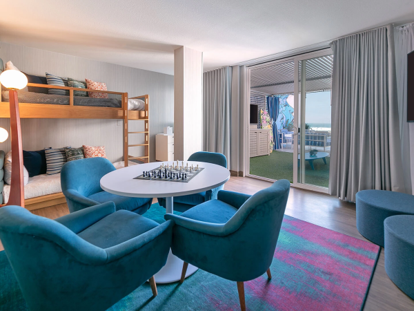 A bright, modern living space with a bunk bed, blue chairs around a round table, colorful rug, floor lamp, and a large TV, plus sliding glass doors to an outdoor patio.