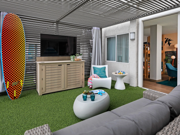 A cozy outdoor lounge with a surfboard, wicker furniture, a TV, planters, a grey sofa, round coffee tables, artificial grass, and sliding doors to an indoor seating area.