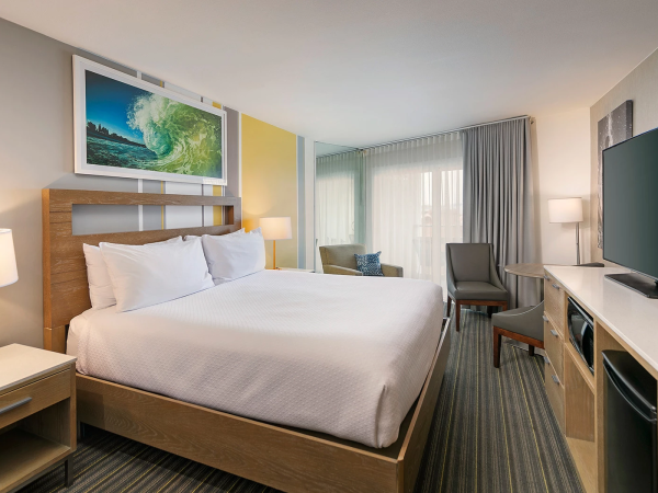 A modern hotel room with a large bed, nightstands, artwork, a desk and chair, a flat-screen TV, and a window with sheer curtains, all in neutral tones.