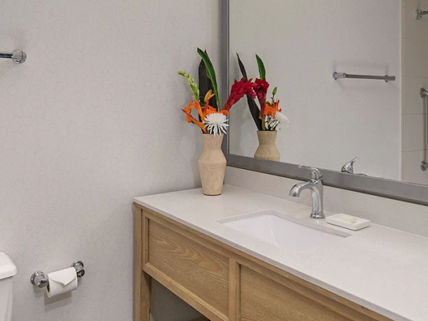 A modern bathroom with a light wooden vanity, white countertop, a single sink, a large mirror, and towels stored on an open shelf; fresh flower arrangements add color.