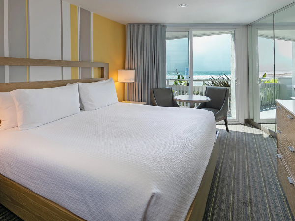 A bright hotel room with a king bed, yellow accent wall, balcony with a seaside view, and a modern dresser and workspace, ending with a period.