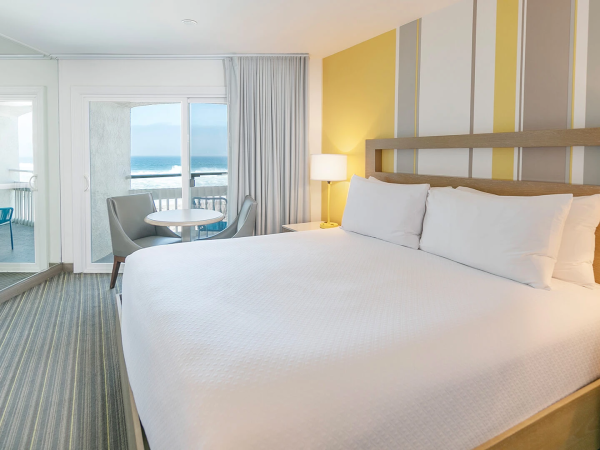 A bright hotel room with a large white bed, nightstands, a seating area by sliding doors to a balcony, and a view of the sea.