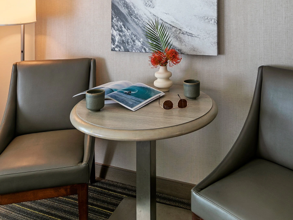 A small two-chair hotel nook with a round table, a lamp, magazines, two mugs, sunglasses, and a decorative flower arrangement on the table.