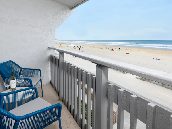 A balcony with two blue chairs and a small table on the left, overlooking a sandy beach and calm ocean under a clear sky.
