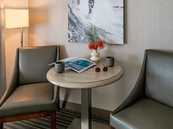 A cozy hotel corner with two gray chairs flanking a small round table, a black-and-white surfing artwork, a lamp, and a vase of flowers on the table.