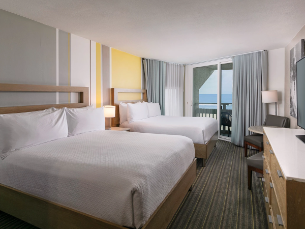 Two large beds in a bright hotel room with a balcony, desk, TV, and coastal view through sliding doors.