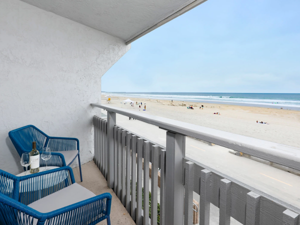 Balcony with two blue chairs and a small table, overlooking a sunny beach with people sunbathing and a calm blue ocean in the distance.