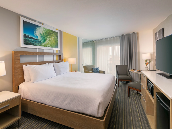 A modern hotel room with a large bed, nightstands, a desk, a chair, a flat-screen TV, and a bright windowed seating area, nicely lit.