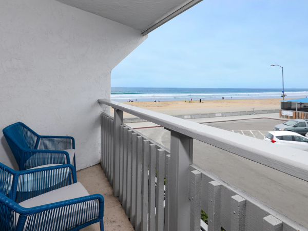 A small balcony with blue wicker chairs overlooks a sunny beach and sea, with a road and parked cars below.