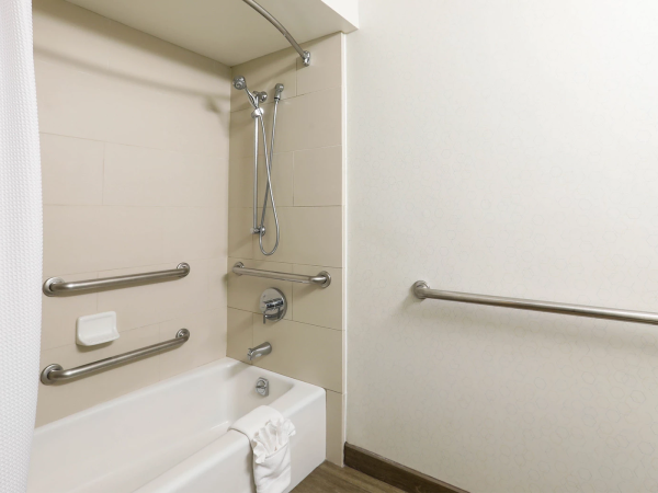 A bathroom with a bathtub/shower combo, grab bars on walls, a handheld showerhead, soap dish, and a towel on the tub edge. It&rsquo;s clean and accessible.