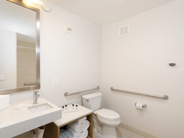A clean, accessible bathroom with a sink, mirror, toilet, towel shelf, grab bars, and folded towels under the sink, all in a bright, white space.