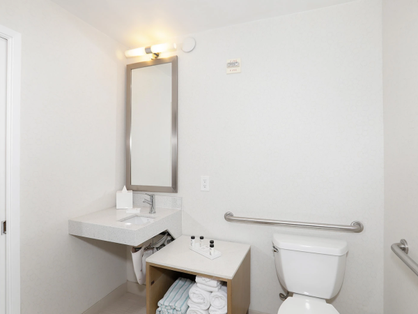 A small, clean bathroom with a wall mirror, light fixture, sink, open shelf with towels, and a toilet with grab bars on the wall. end.