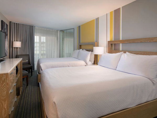 A modern hotel room with two queen beds, a desk, and a TV, bright and tidy with neutral tones and a colorful accent wall.