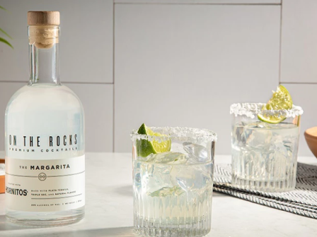 A bottle of On The Rocks premium cocktail mix with two prepared margarita-style drinks on a white marble counter, lime garnish, and a bowl of limes in the background.