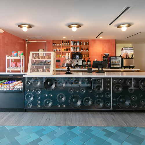 A modern cafe bar with a long counter, glass display, and large speaker-woven front, set in a bright, inviting space with shelves behind the bar, coffee gear, and drinks, ending with a period.