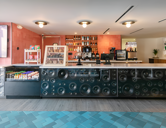 A modern cafe bar with a long counter, glass display, and large speaker-woven front, set in a bright, inviting space with shelves behind the bar, coffee gear, and drinks, ending with a period.