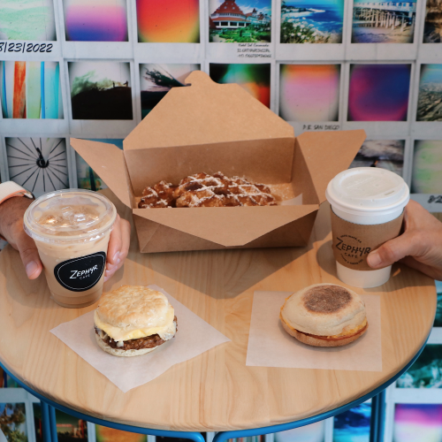 Two people share a round table with coffee drinks and pastries: a donut, a muffin, and a boxed pastry, against a colorful tiled backdrop.