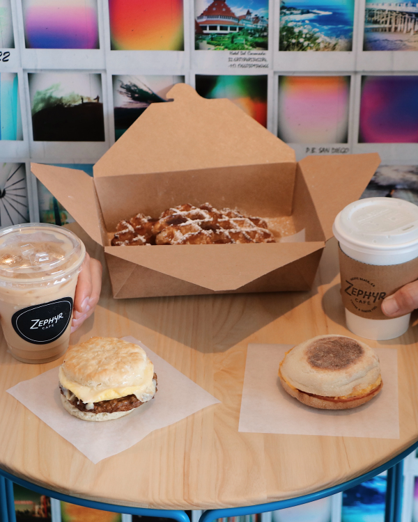 Two people share a round table with coffee drinks and pastries: a donut, a muffin, and a boxed pastry, against a colorful tiled backdrop.