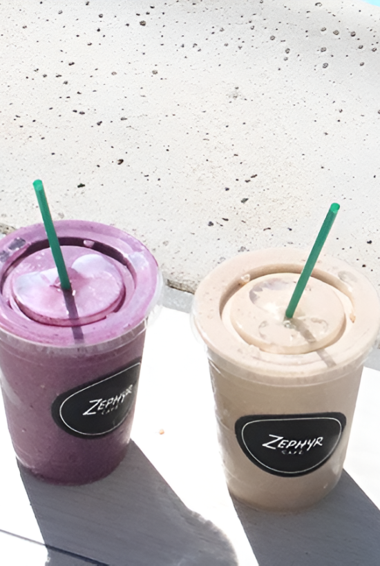 Two iced beverages with green straws sit on a white ledge near a pool, one purple and one beige, both in takeaway cups.