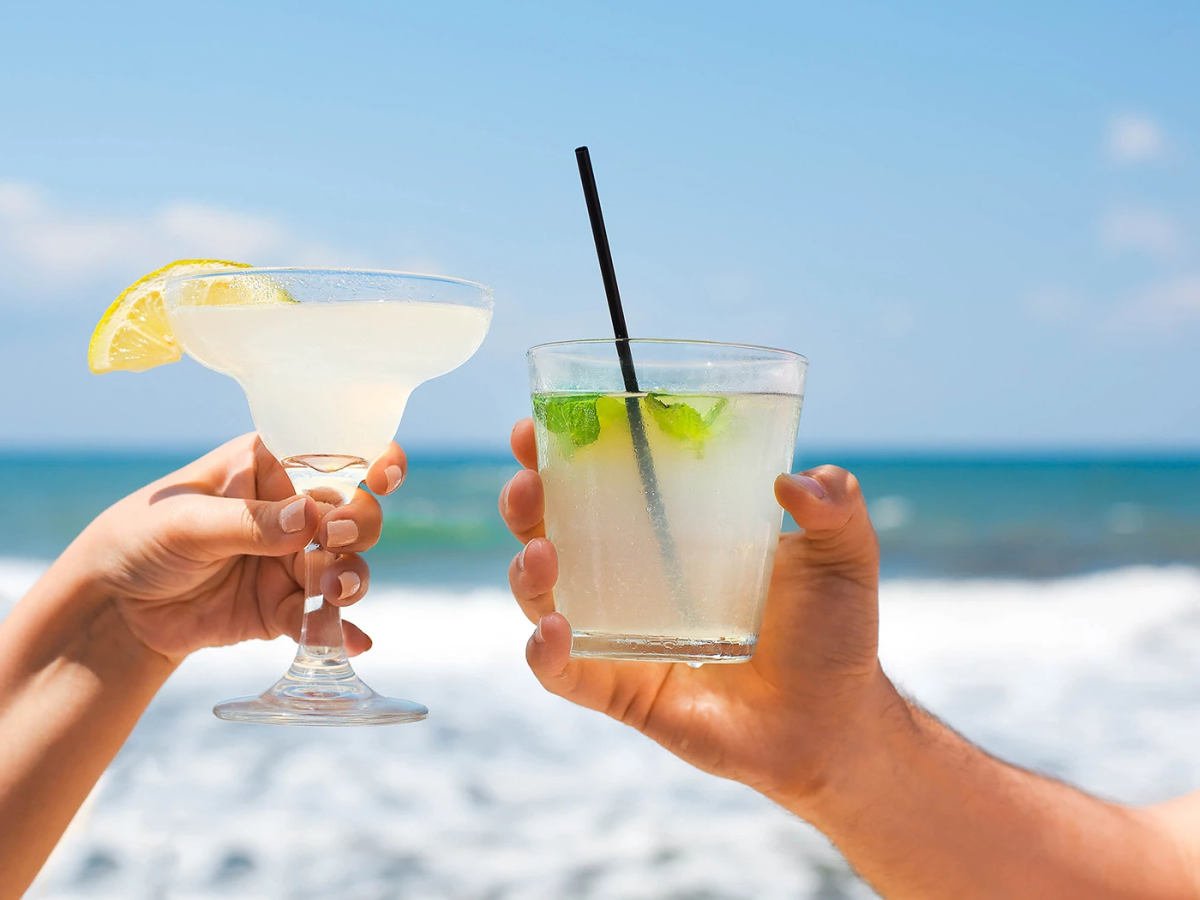 Two hands raise cocktails in a beach scene, clinking glasses with lime and lemon slices against a sunny ocean backdrop.