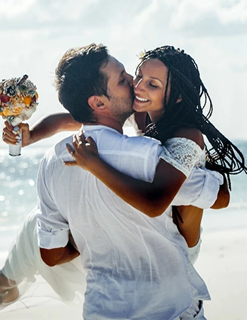 A couple at the beach, the groom lifts the bride in a kiss, she holds a bouquet, ocean waves sparkle in the background.