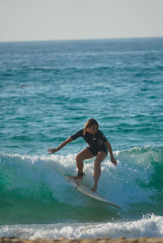 A person surfing a small wave along a sandy beach, balancing low, knee-bent stance as the turquoise water splashes.