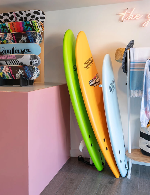 Surf boards and racks in a bright shop: stacked boards on a pink counter, bright neon sign, and a rack with wetsuits and gear.
