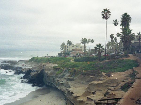 Coastal cliff with a wooden railing, rocky shoreline, palm trees, and distant houses along a sea-facing cliff under a cloudy sky.