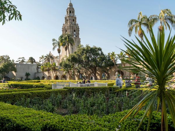 A grand, ornate church tower rises behind a manicured garden with palm trees and hedges, sunny sky overhead.
