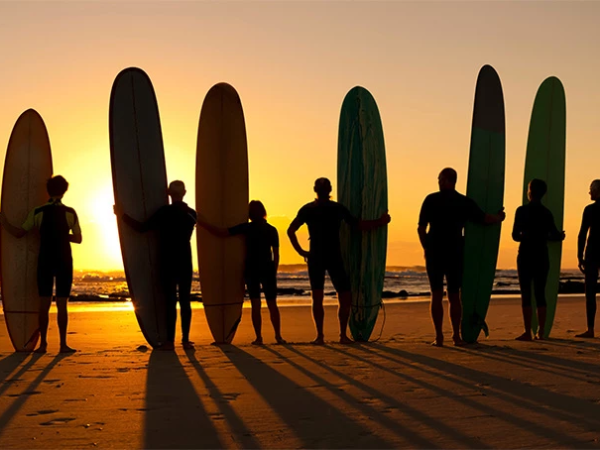 A group of surfers with their boards stand in a line on a sunset beach, long shadows stretching toward the camera.