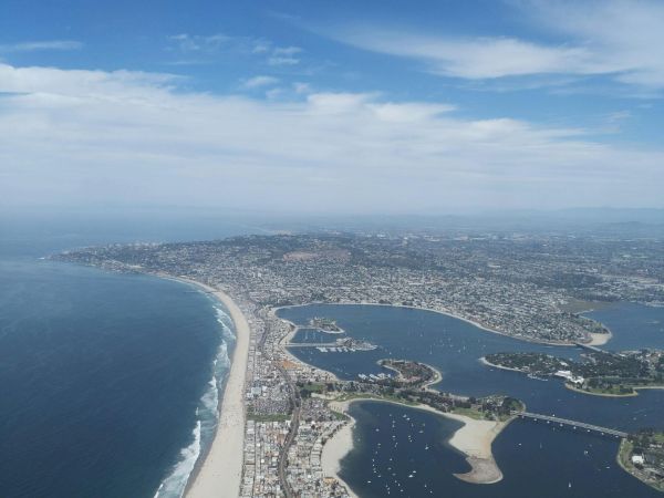 Aerial view of a coastal city with a long sandy beach, curved shoreline, and waterways forming lagoons and inlets along the coast, bright blue sky, and calm seas.