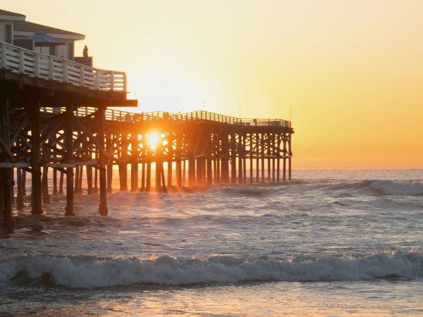 A wooden pier extends into the ocean at sunset, with warm light glowing through the gaps and gentle waves rolling in, calm seaside scene.